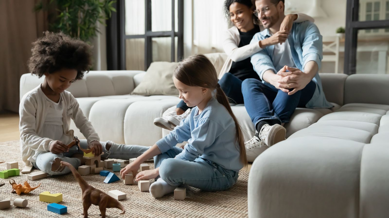 Two children playing with blocks on a living-room floor while their parents sit together on the sofa behind them.