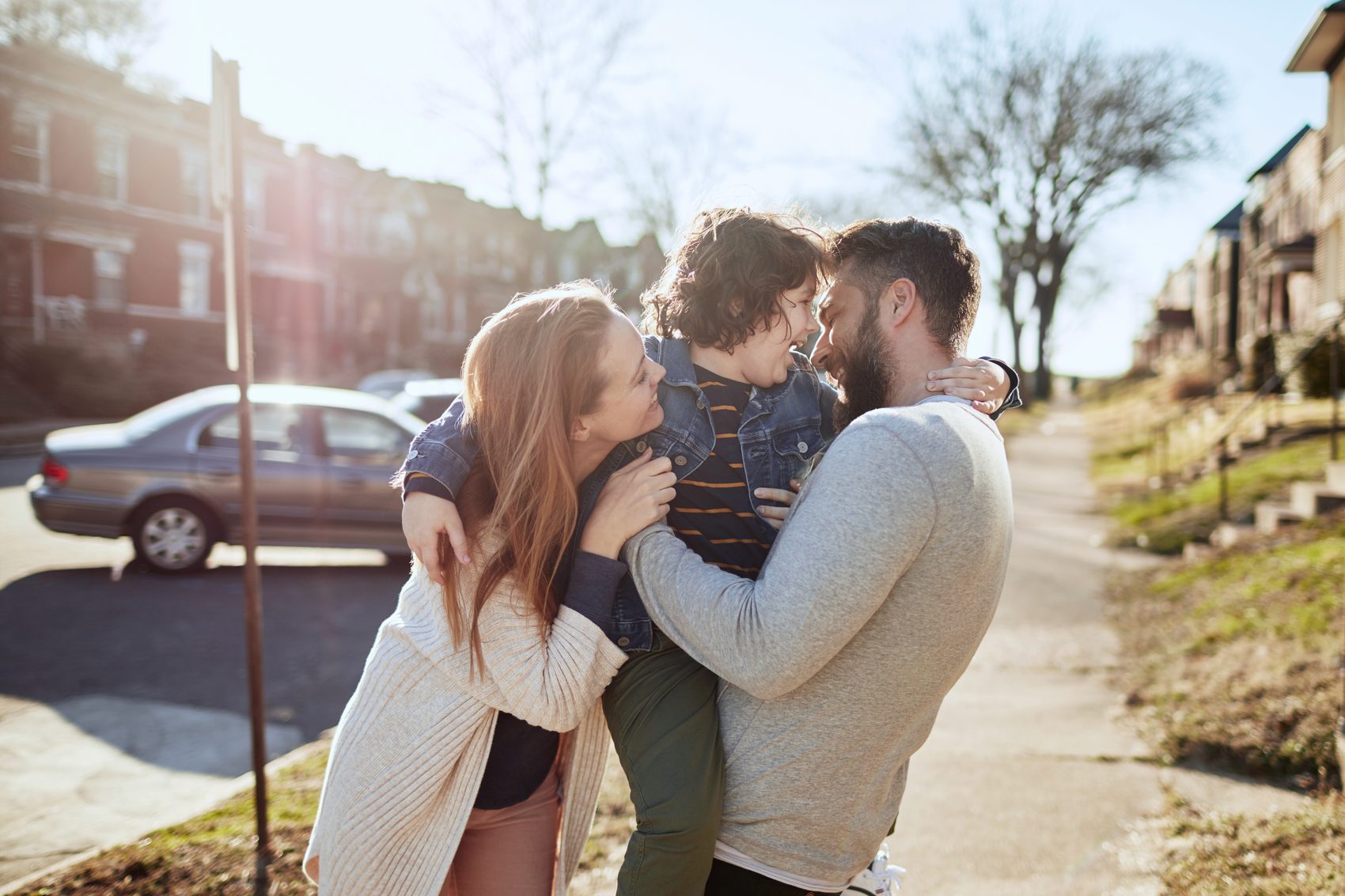 Parents on a sunlit street, the father holding their small child between them, foreheads touching.